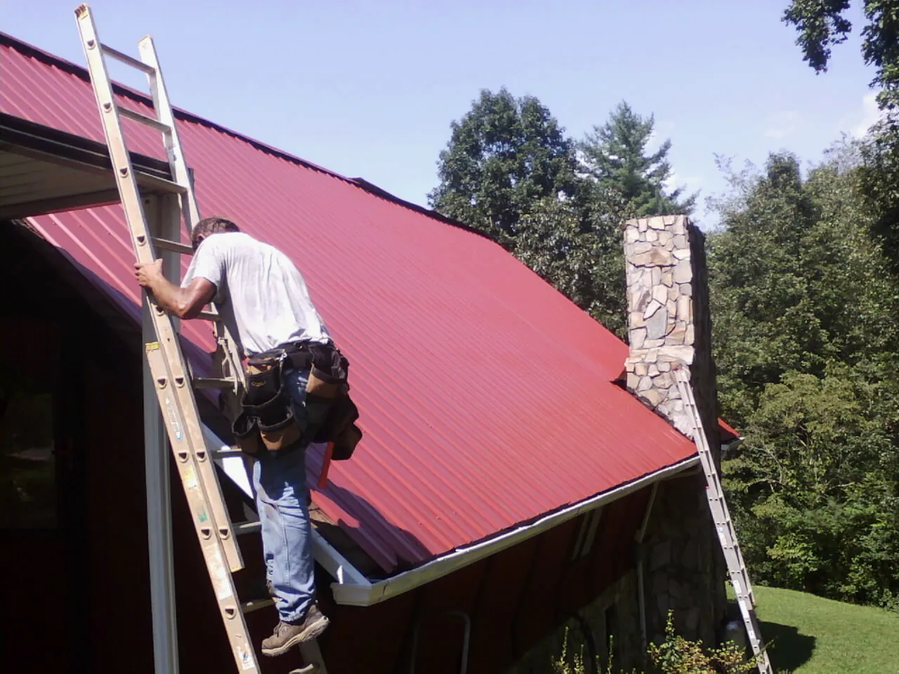 Roofer on a ladder beside a red metal roof and stone chimney — Soffit & Fascia Repair by Roofing Brothers in Nunica